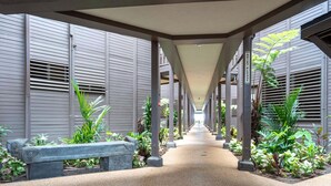 A covered walkway with benches and plants, leading to a building with grey siding. - Hale Hui Kai 208 - Ocean View Luxury Condo on the Best Beach on Maui, Split AC! (Kihei)