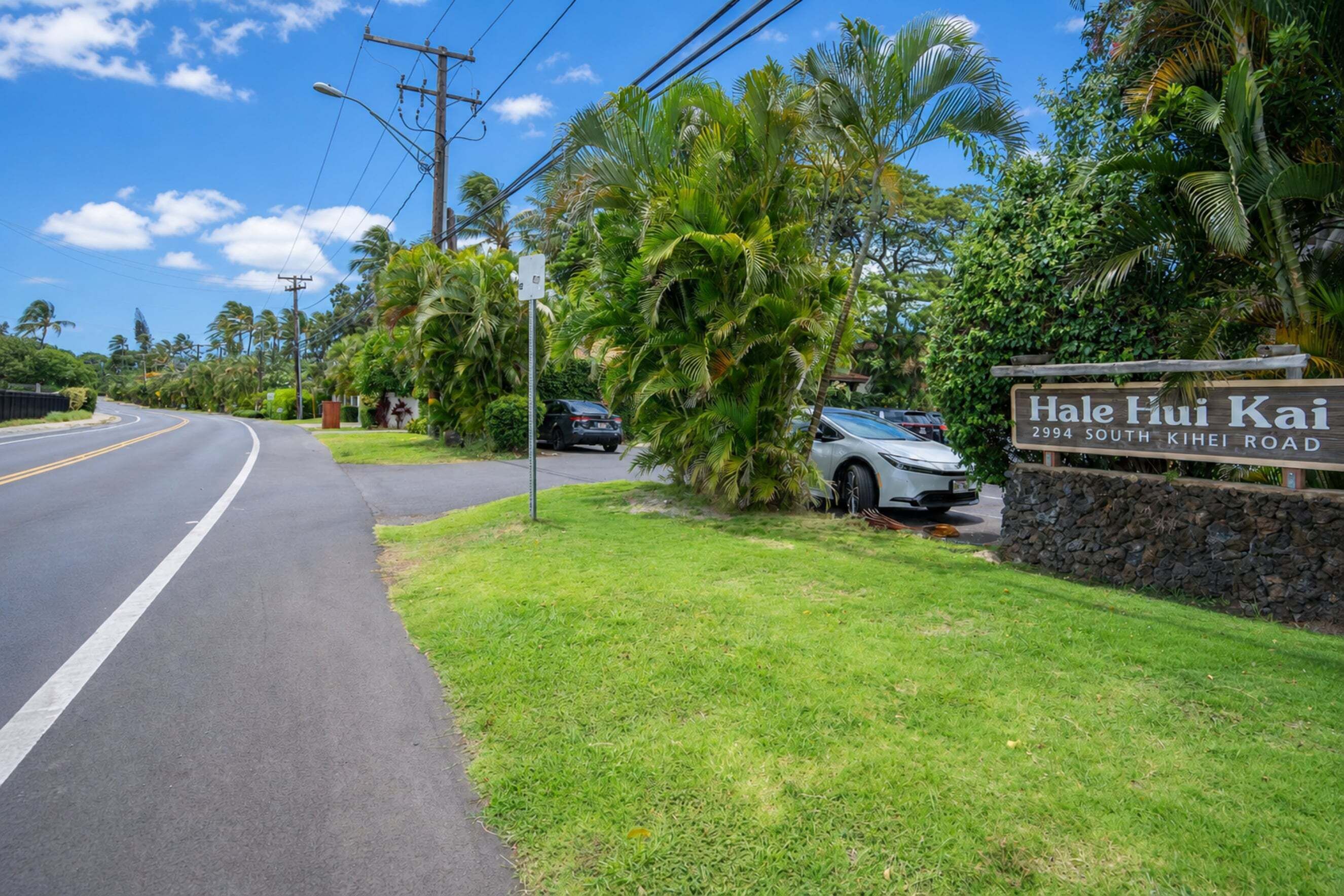 A street view with a sign for Hale Fui Kai on South Kihei Road, surrounded by lush greenery and palm trees.