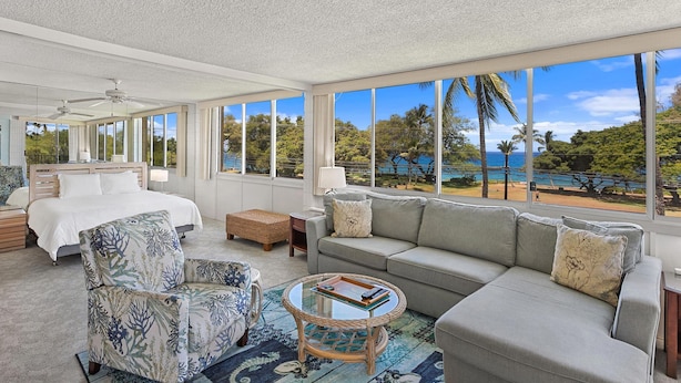A spacious room with a large bed, a grey sofa, a wicker coffee table, and a view of the beach and ocean. - Haleakala Shores A-301 - Golden Sand Beaches, Ocean Views, Sleeps 6, Split AC! (Kihei)