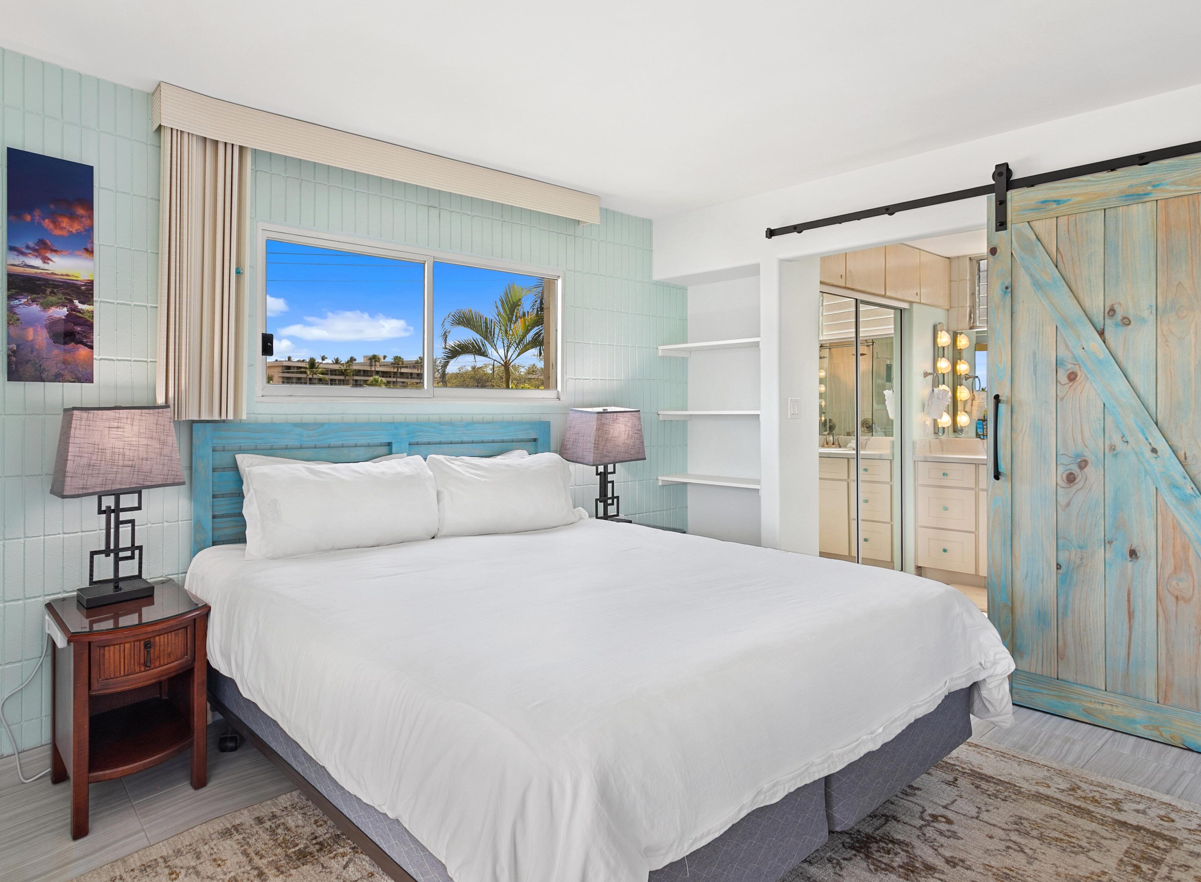 A bedroom with a large bed, a nightstand, a window with a view of palm trees, and a wooden wardrobe with a sliding door.