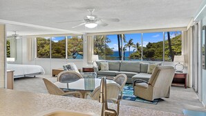 A modern hotel room with a large window offering a view of the ocean and palm trees. There is a dining area with a table and chairs, a sofa, and a ceiling fan. - Haleakala Shores A-301 - Golden Sand Beaches, Ocean Views, Sleeps 6, Split AC! (Kihei)