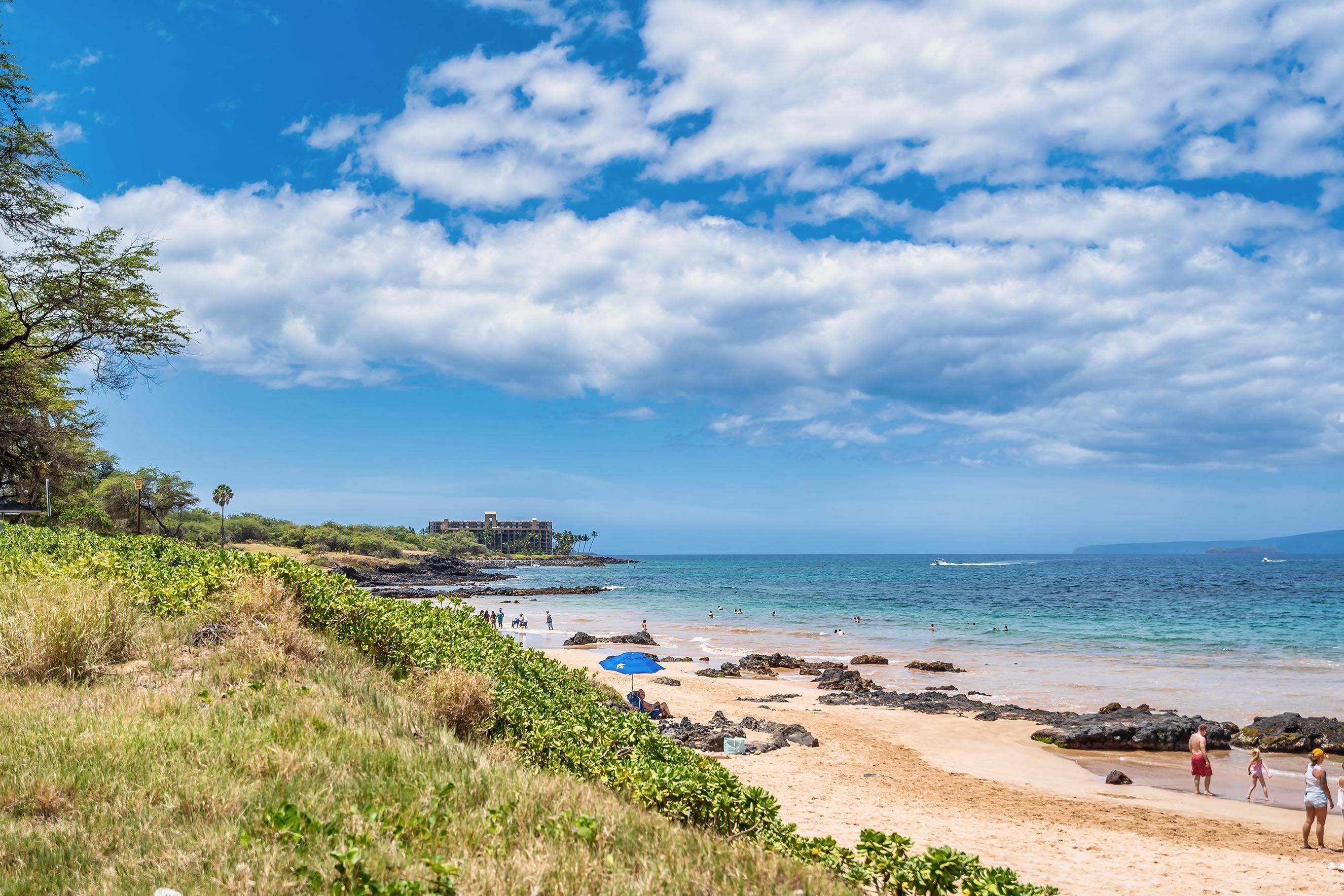 A beach with clear skies, rocky shoreline, and a building in the background.