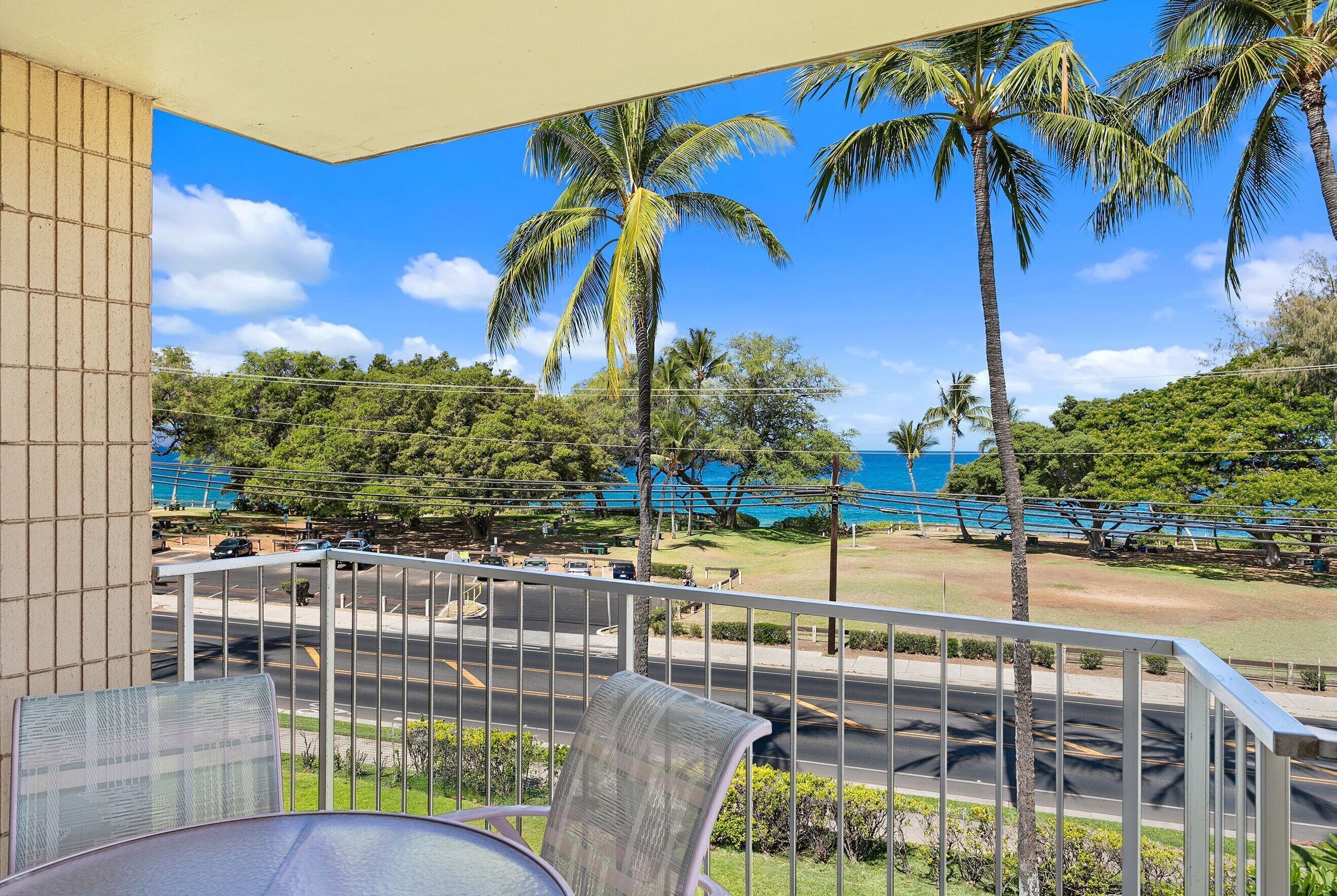 A balcony with a view of the ocean, palm trees, and a road with vehicles.