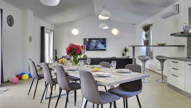 Un comedor moderno con una larga mesa dispuesta para seis personas, rodeada de sillas. Se observa una zona de cocina con estufa y fregadero a la derecha.
