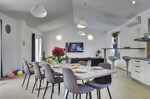 Ein moderner Essbereich mit einem langen Tisch, gedeckt für sechs Personen, umgeben von Stühlen. Eine Küchenzeile mit Herd und Spüle ist rechts zu sehen.