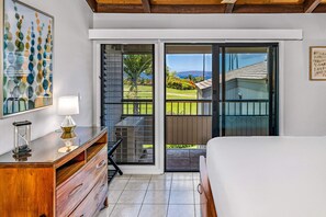 Ein Schlafzimmer mit einem Bett, einer Kommode, einer Lampe und einem großen Fenster mit Blick auf einen Garten und ein Gebäude.