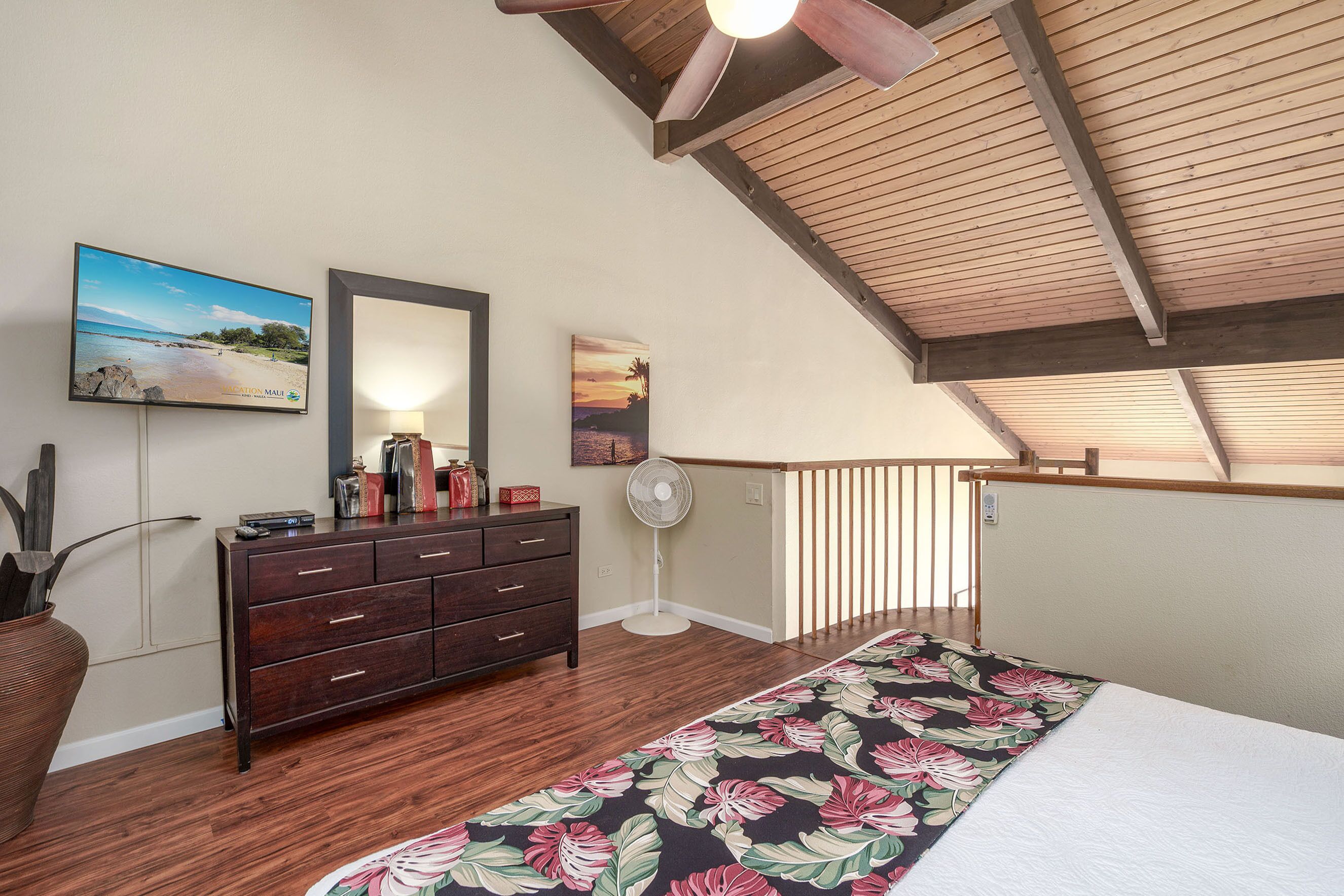 Une chambre avec un plafond en bois, un grand lit recouvert d’une couvre-lit à motifs floraux, une commode, un téléviseur fixé au mur et un ventilateur.