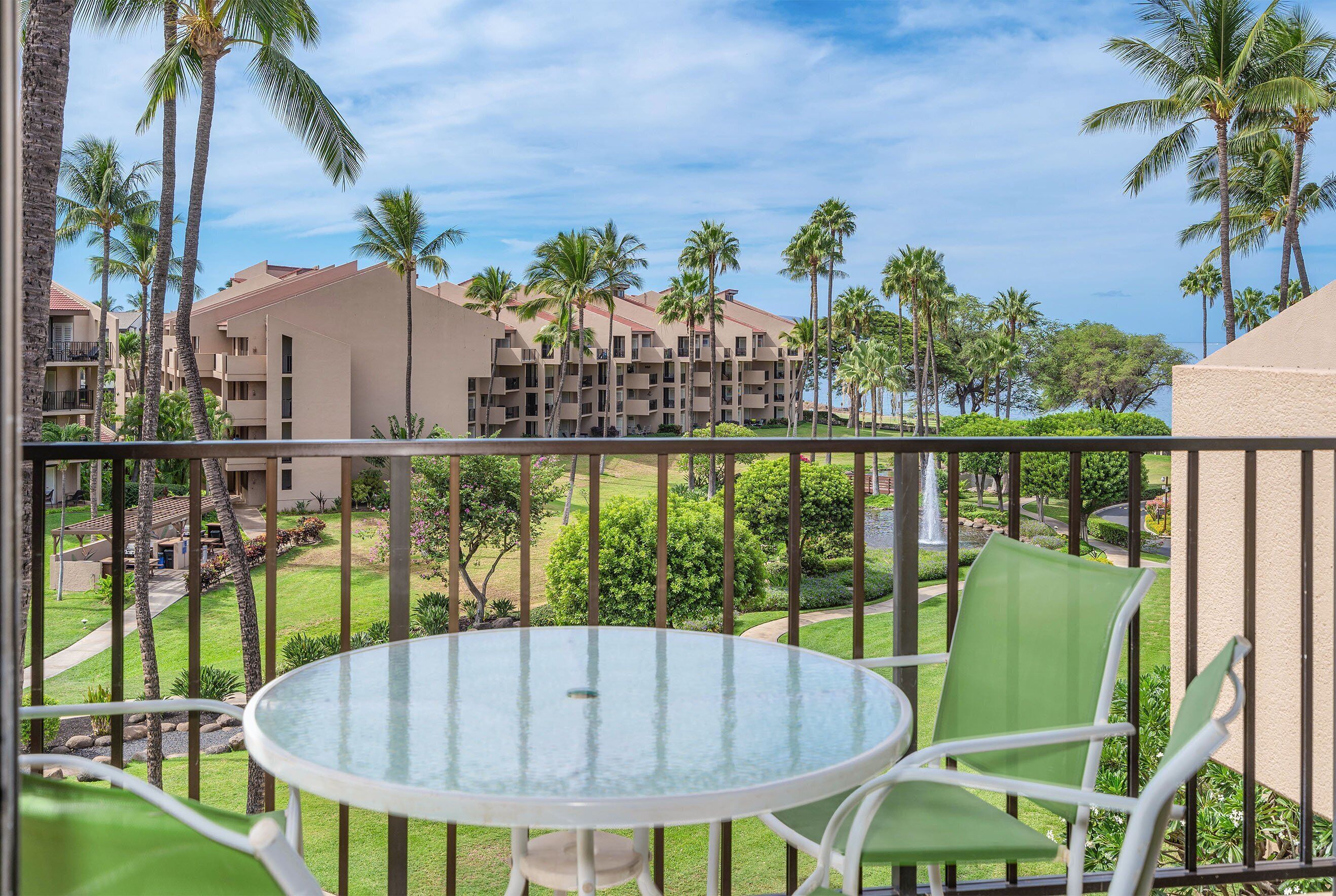 A balcony with a glass table and chairs, overlooking a courtyard with palm trees and a building with multiple balconies.