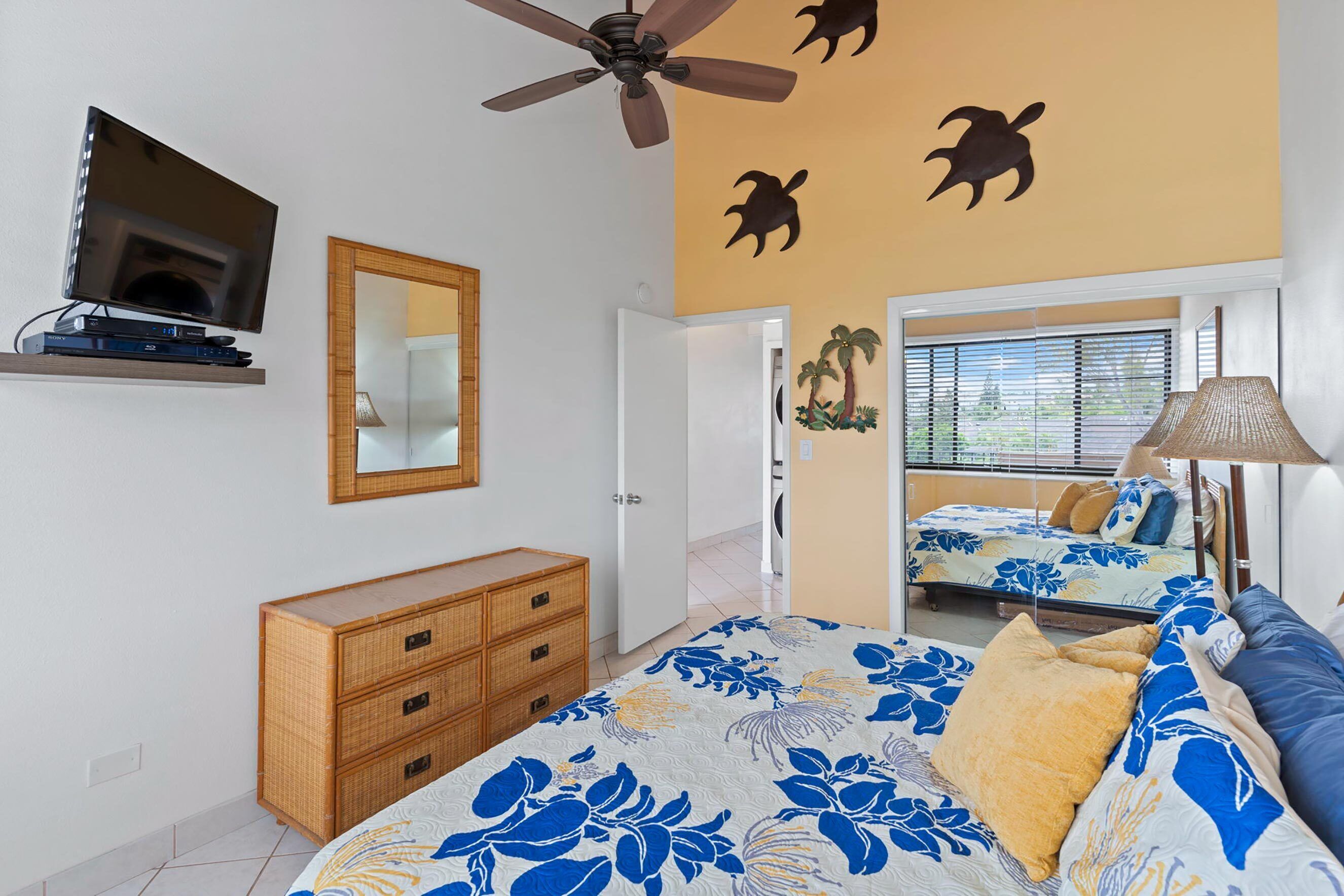 A bedroom with a bed, a television mounted on the wall, a ceiling fan, and a mirror.