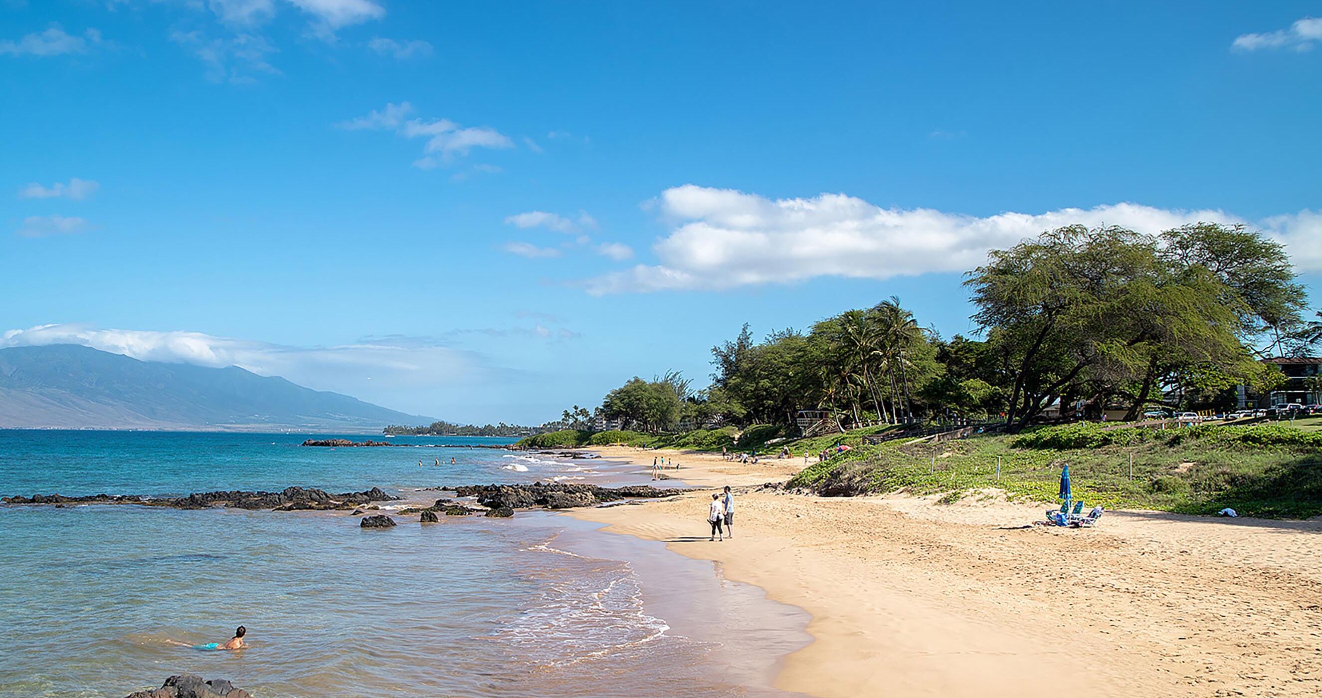 A beach with clear blue water, a sandy shore, and a few people walking.