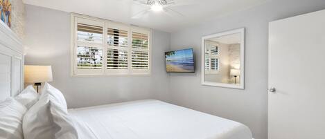 A bedroom with a bed, a ceiling fan, a window with blinds, a painting, and a door.