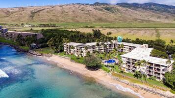 A coastal hotel with multiple buildings, a beach, and wind turbines in the background.