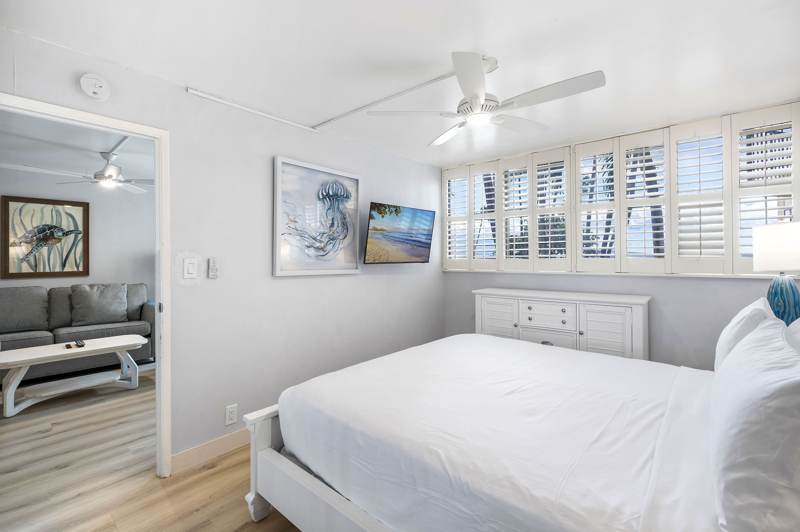 A bedroom with a bed, a ceiling fan, a window with blinds, a bedside table, and a wall-mounted TV.