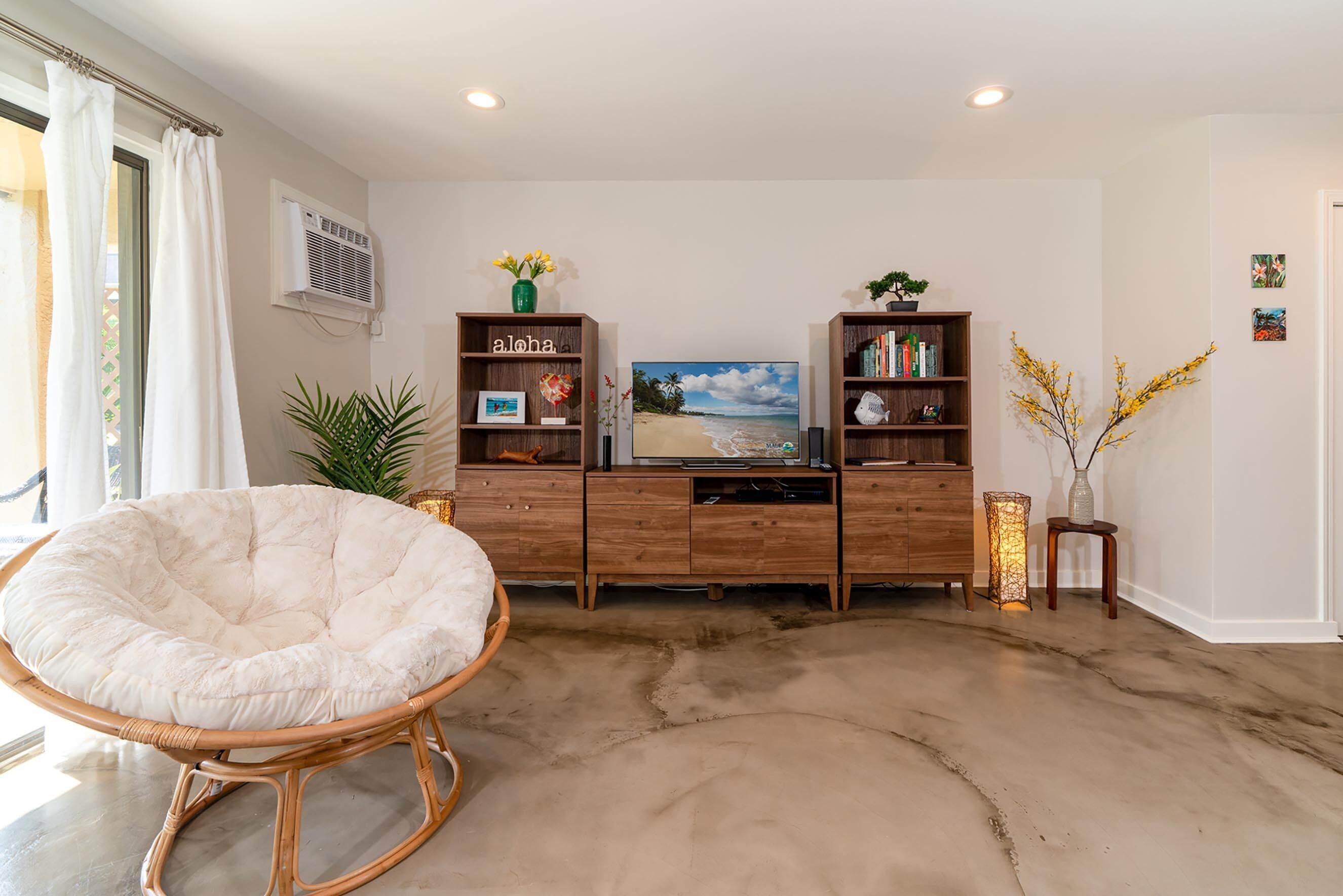 A living room with a large white cushioned chair, wooden bookshelves, a flat-screen TV, and a beach-themed painting.