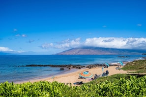 A beach with clear blue water, a sandy shore, and a mountain in the background. There are umbrellas and chairs on the beach. - Kamaole Sands 4-306 (Kihei)