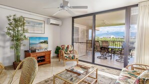 A spacious living area with a glass coffee table, wicker furniture, a sofa with a tropical print, a television, and a large window offering a view of a balcony and mountains. - Kamaole Sands 4-306 (Kihei)