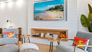 A modern living room with a beach-themed TV, comfortable seating, and a wooden shelf with books and remote controls. - Summer Sale! Deluxe Modern Renovation, Split-ac, Kamaole Sands 4-411, Book Now! (Kihei)