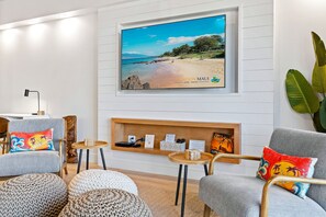 A modern living room with a beach-themed TV, comfortable seating, and a wooden shelf with books and remote controls.
