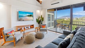A modern living room with a large TV, comfortable seating, a ceiling fan, and a balcony with a view. - Summer Sale! Deluxe Modern Renovation, Split-ac, Kamaole Sands 4-411, Book Now! (Kihei)