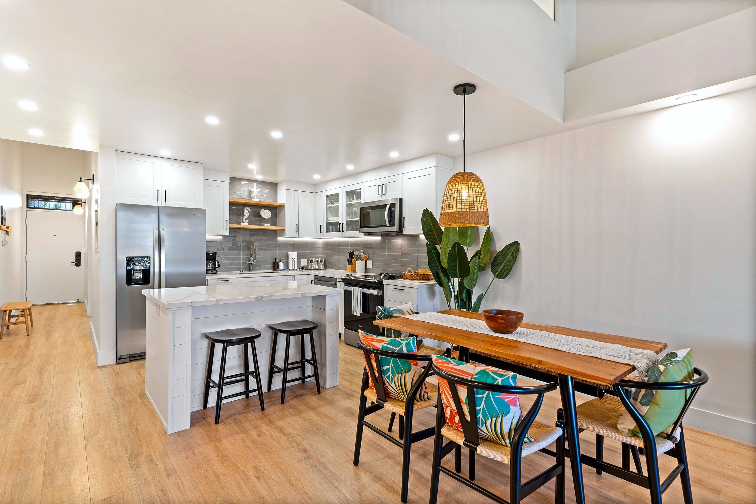 A modern kitchen with white cabinetry, a central island with stools, and a dining area with a wooden table and colorful chairs.