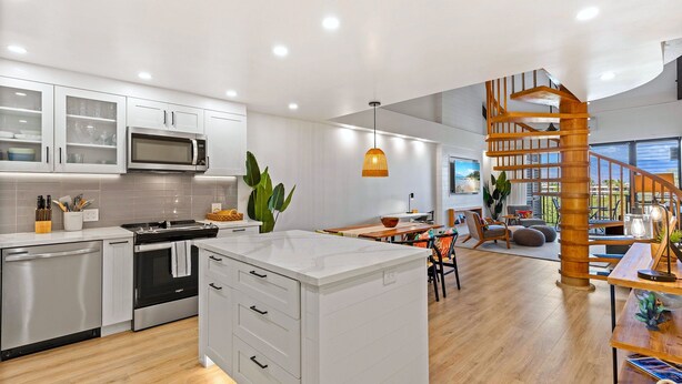 A modern kitchen with white cabinetry, a central island, and a dining area with a wooden staircase. - Summer Sale! Deluxe Modern Renovation, Split-ac, Kamaole Sands 4-411, Book Now! (Kihei)