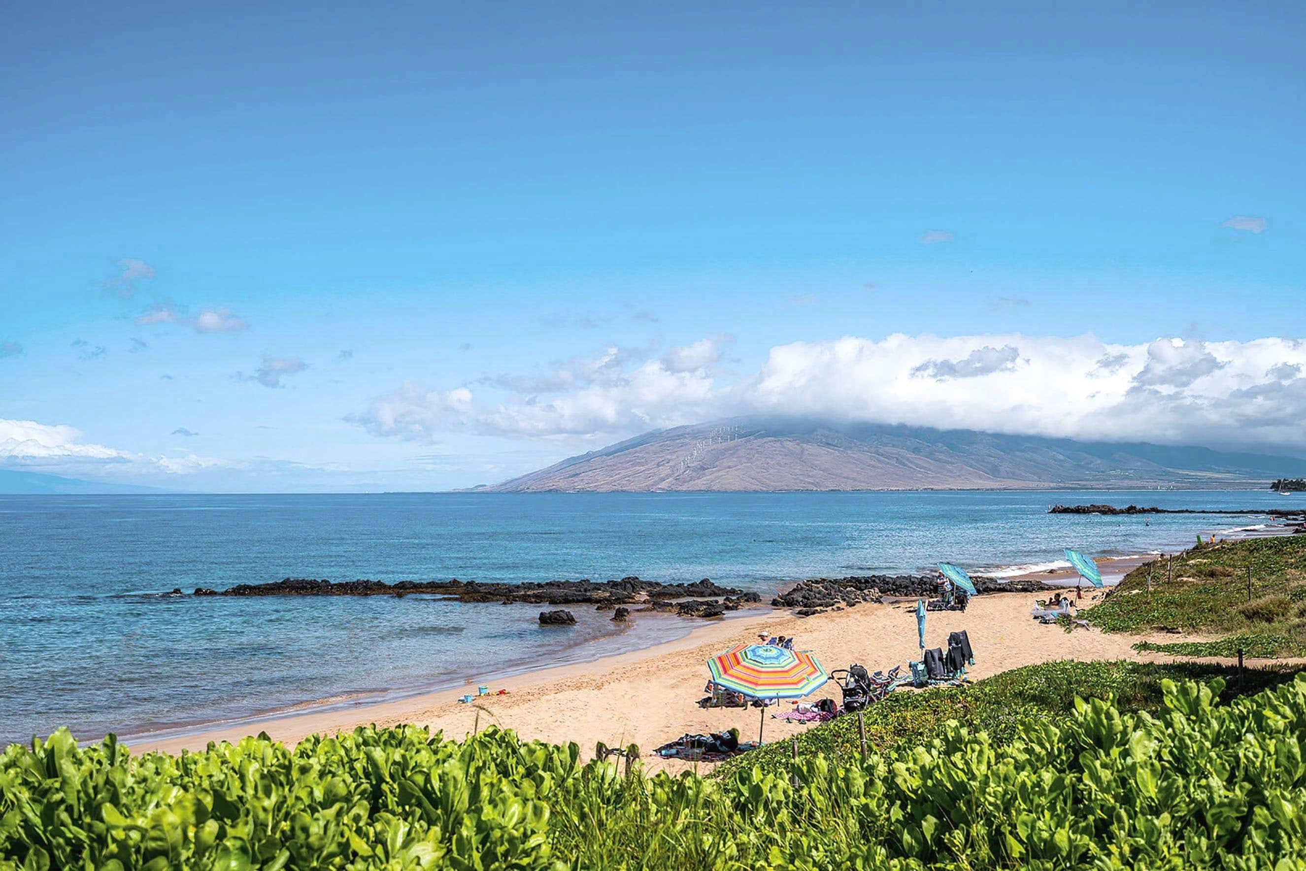 A beach with a clear view of the ocean, a mountain in the distance, and a rocky shoreline.