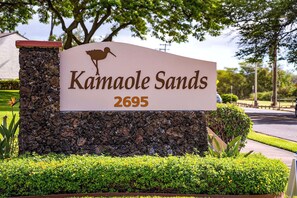A sign for Kamaole Sands with the address 2695, set against a backdrop of landscaped greenery and trees.