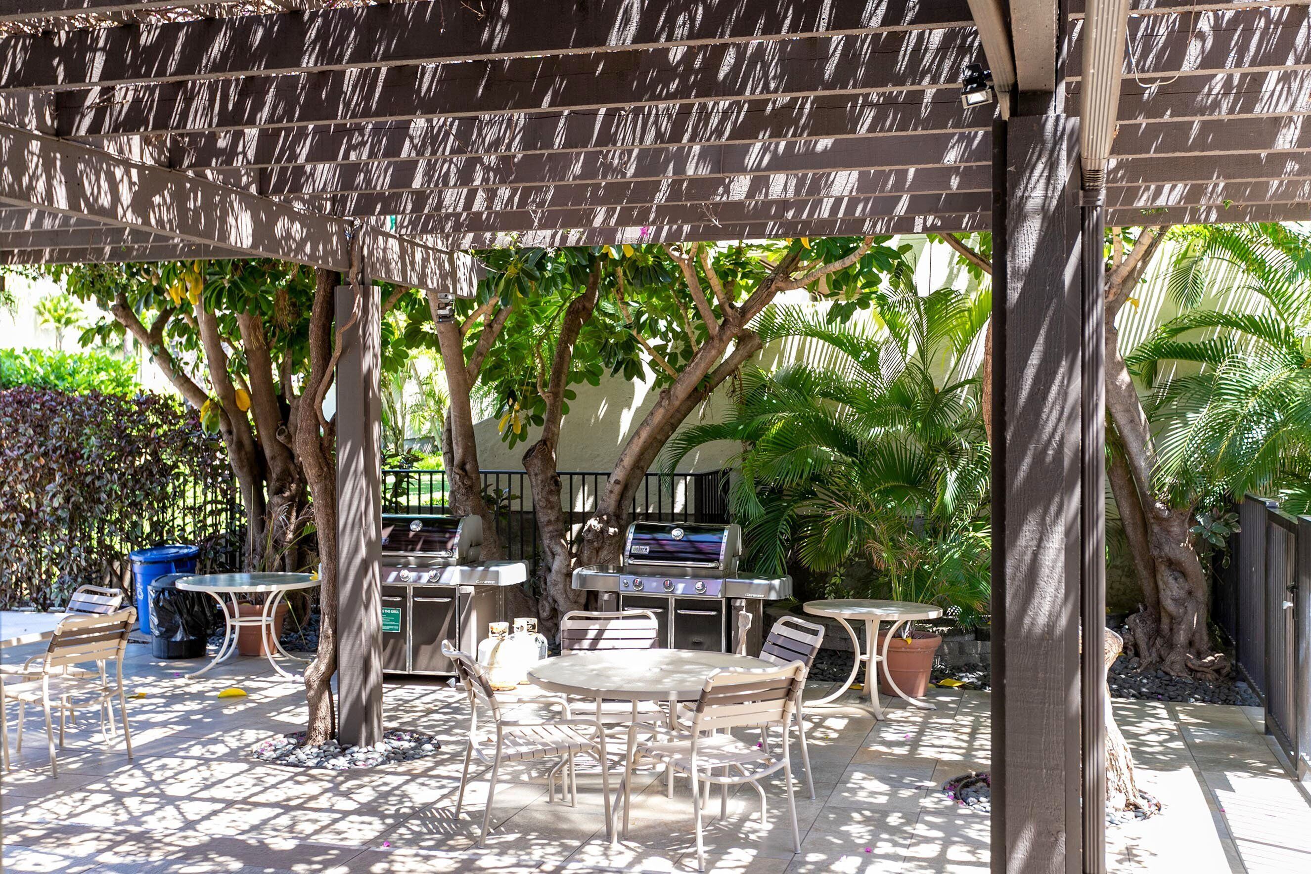 An outdoor patio with a grill, tables, and chairs, surrounded by trees and plants.