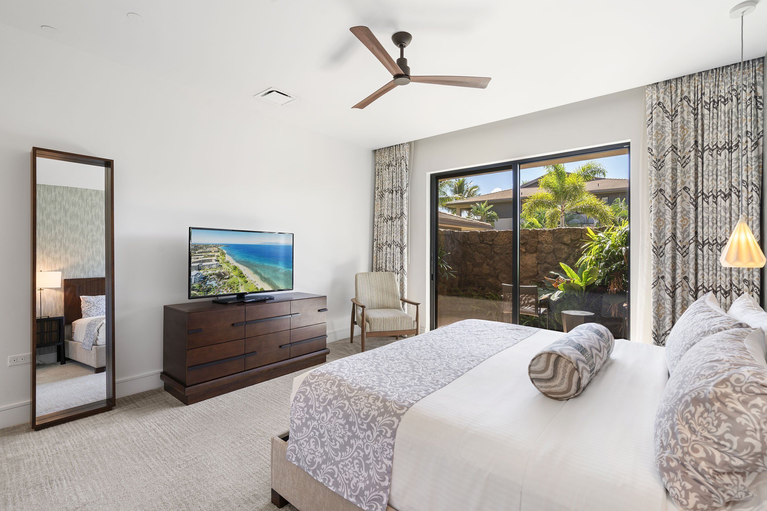 A bedroom with a bed, a television, a ceiling fan, a chair, and a view of the outdoors.