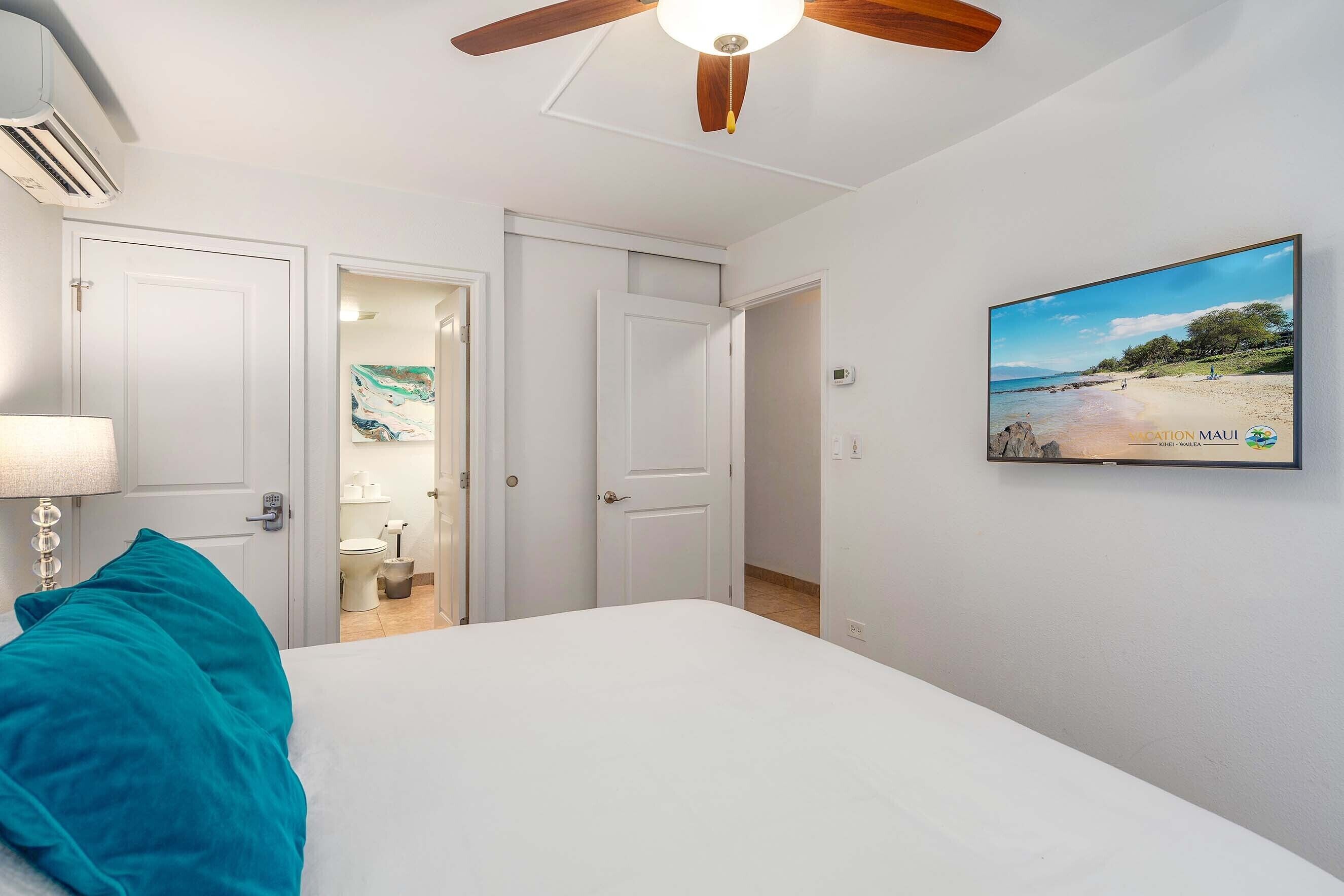 A bedroom with a bed, a ceiling fan, a TV on the wall, and a bathroom visible through an open door.