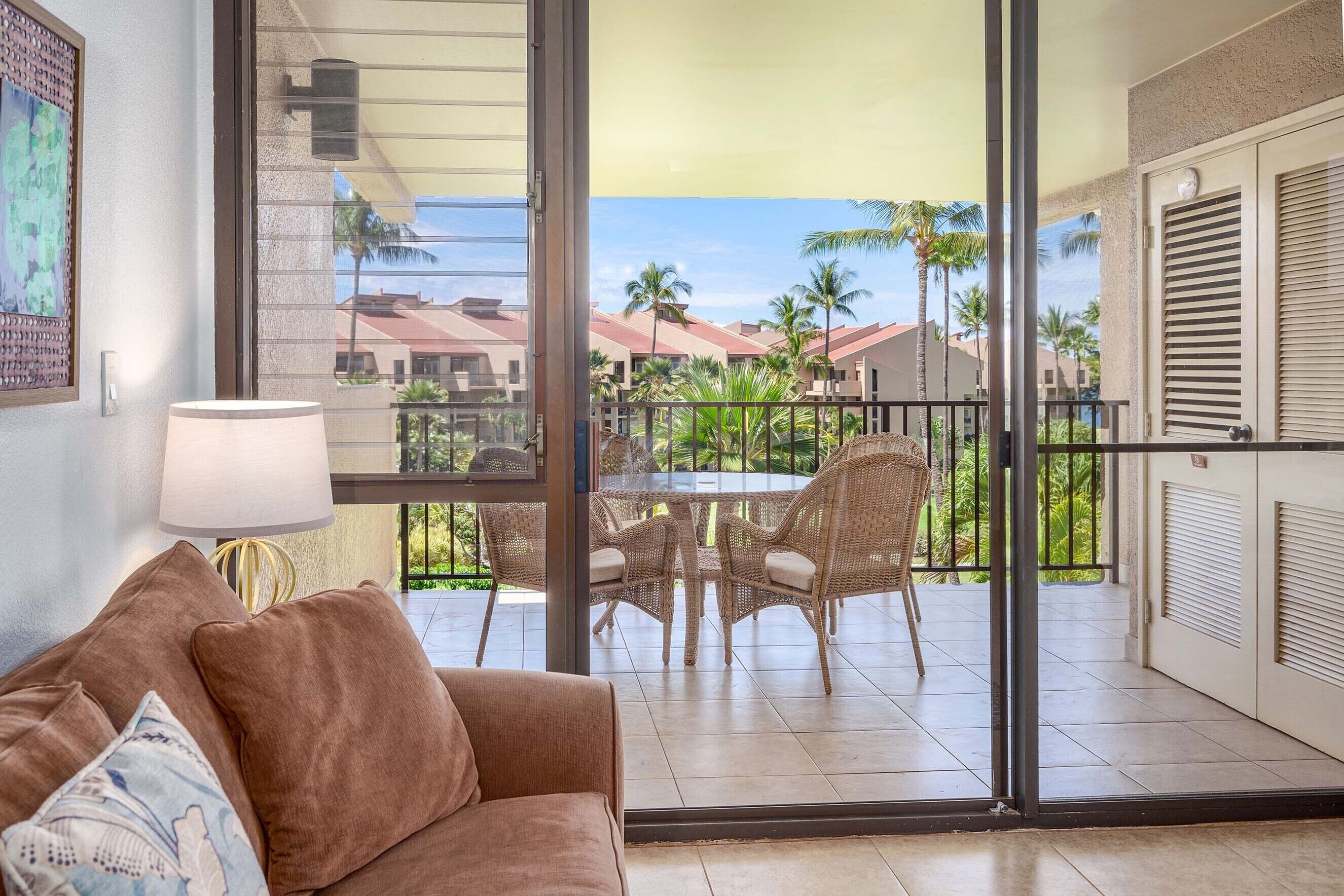 A balcony with wicker furniture, a glass door, and a view of palm trees and buildings.