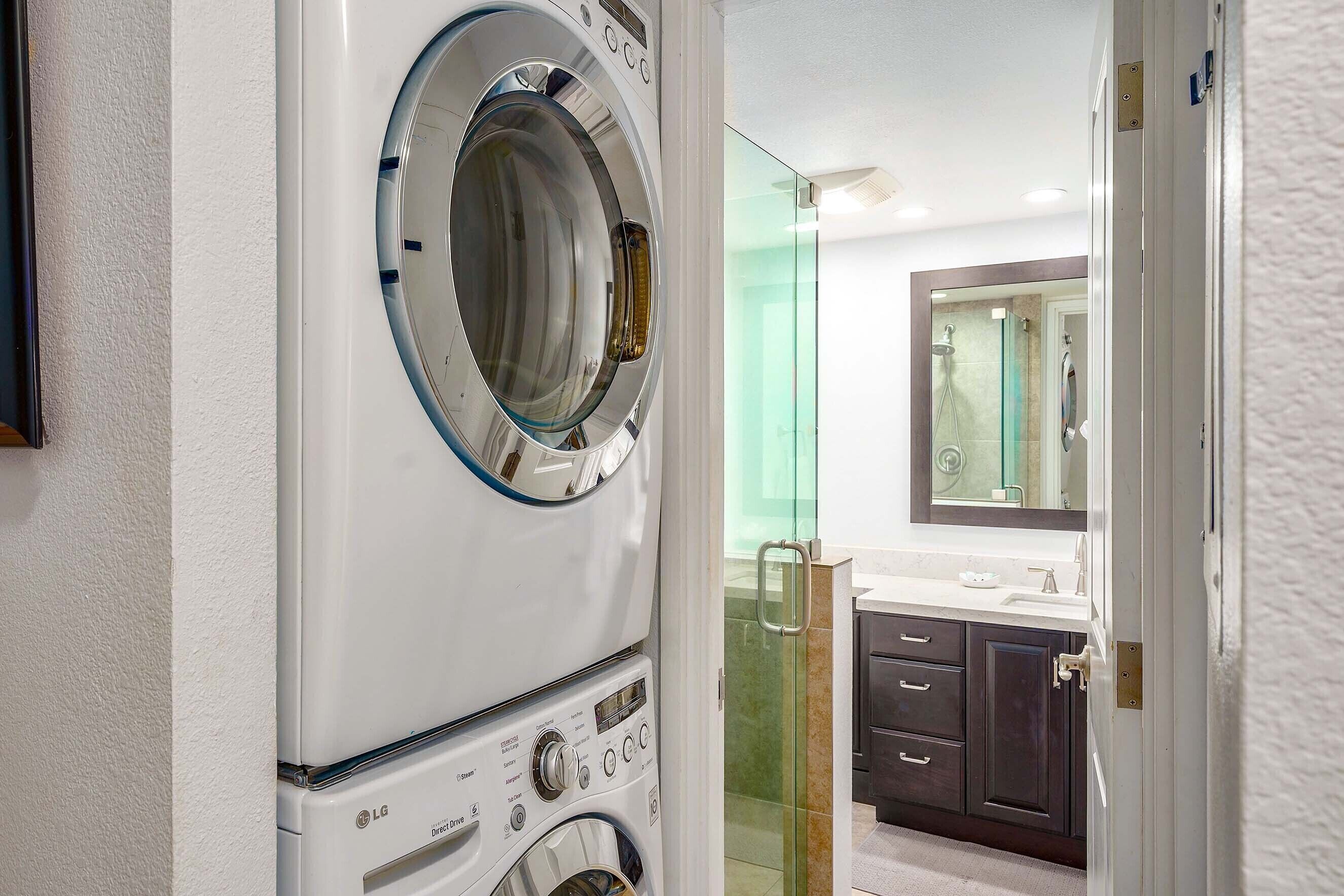 A modern laundry room with a front-loading washer and dryer, a glass shower enclosure, and a bathroom with dark cabinets and a mirror.