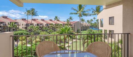 A balcony with wicker furniture and a glass table, overlooking a resort with multiple buildings and palm trees.