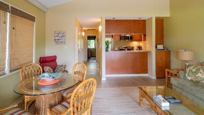 A living room with a glass dining table, wicker chairs, a sofa, and a kitchen area with wooden cabinets and appliances. - Kihei Garden Estates C-201, Ocean Views, Near the Beach, Renovated, w/ Split AC! (Kihei)