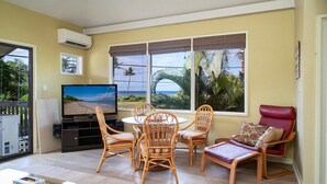 A living room with a TV, wicker furniture, a red chair, and large windows with a view of palm trees and the ocean. - Kihei Garden Estates C-201, Ocean Views, Near the Beach, Renovated, w/ Split AC! (Kihei)