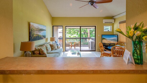 A living room with a sofa, coffee table, and a TV. There is a balcony with a table and chairs. - Kihei Garden Estates C-201, Ocean Views, Near the Beach, Renovated, w/ Split AC! (Kihei)