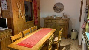 Une table à manger en bois avec des chaises, un buffet en bois, une fenêtre avec des rideaux, une horloge murale et une petite poubelle.