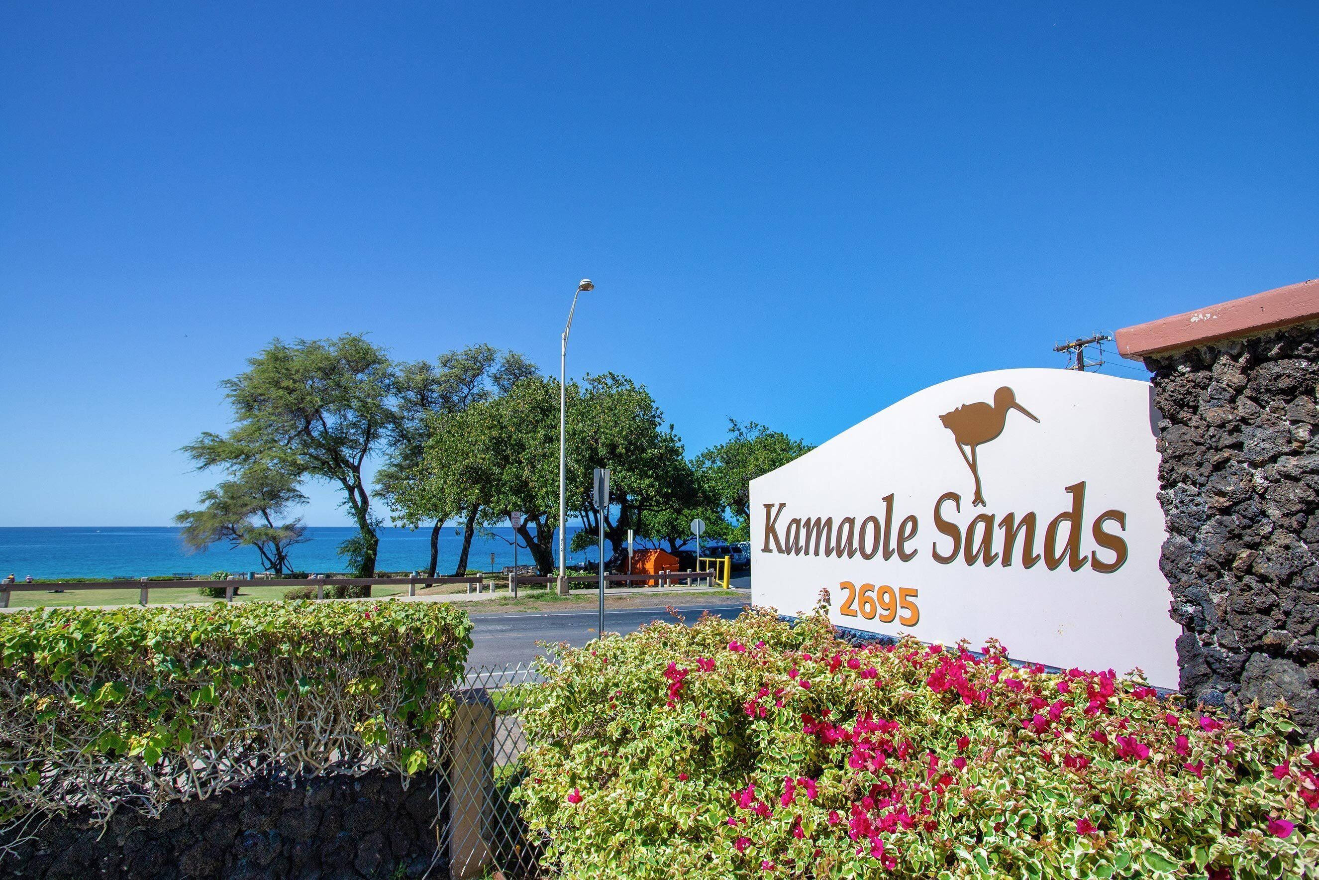 Placa com a inscrição 'Kamaole Sands' e o número 2695, perto de uma praia com árvores e céu claro.