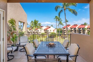 A balcony with a table and chairs, overlooking a resort with palm trees and buildings. - Kamaole Sands 7-405 - Stunning Renovation, Large Lanai, Ocean Views, Split AC! (Kihei)
