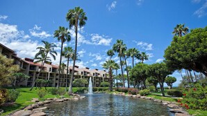 A hotel courtyard with a central pond, palm trees, and a building with multiple balconies. - Kamaole Sands 7-405 - Stunning Renovation, Large Lanai, Ocean Views, Split AC! (Kihei)