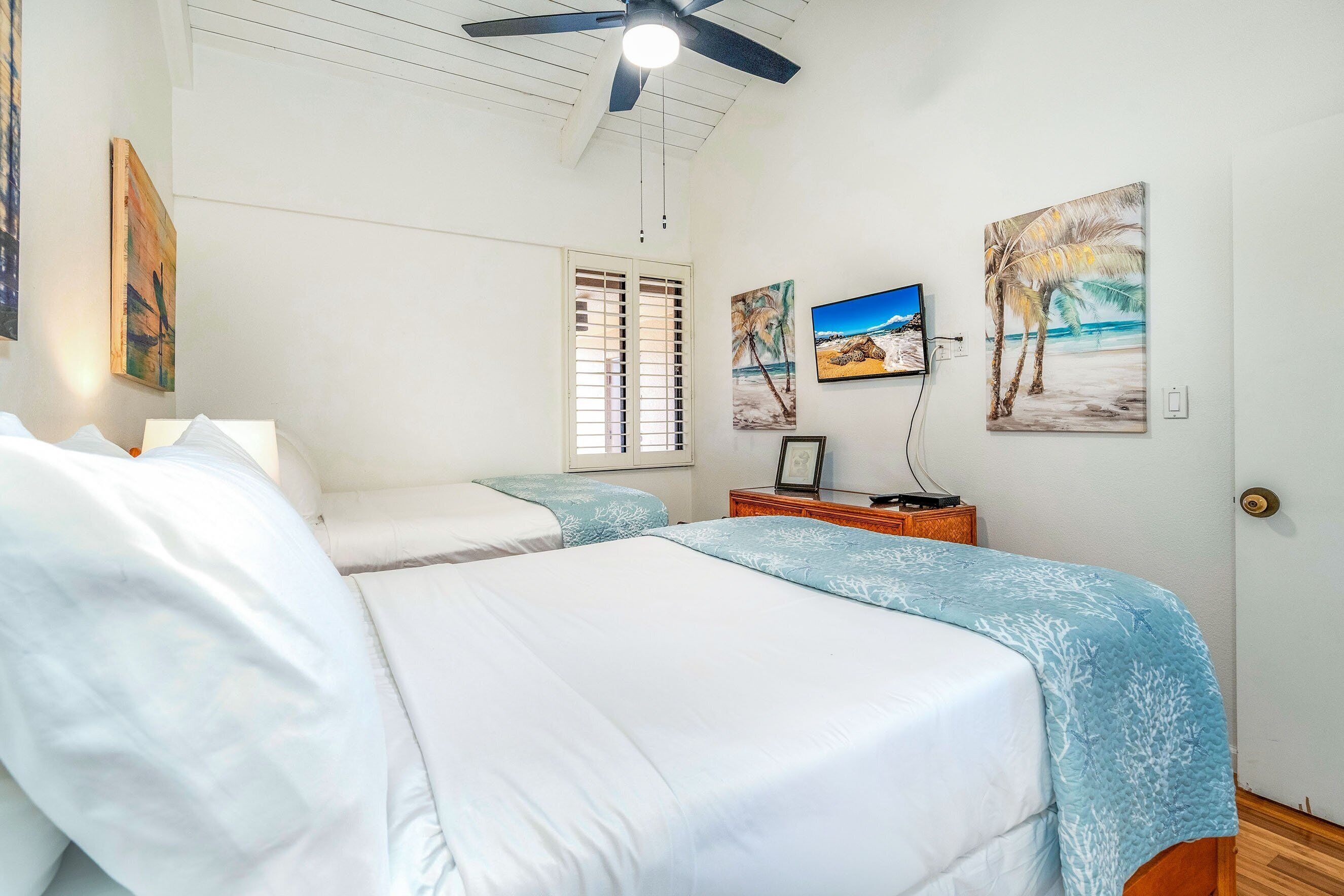 A bedroom with two beds, a ceiling fan, a nightstand, and wall art depicting beach scenes.