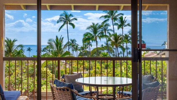 A balcony with a round table and wicker chairs, overlooking a tropical landscape with palm trees and the ocean. - Wailea Ekahi 12D - Budget Friendly Ocean View Studio w/ Lanai, Kitchen, WD, & AC (Kihei)