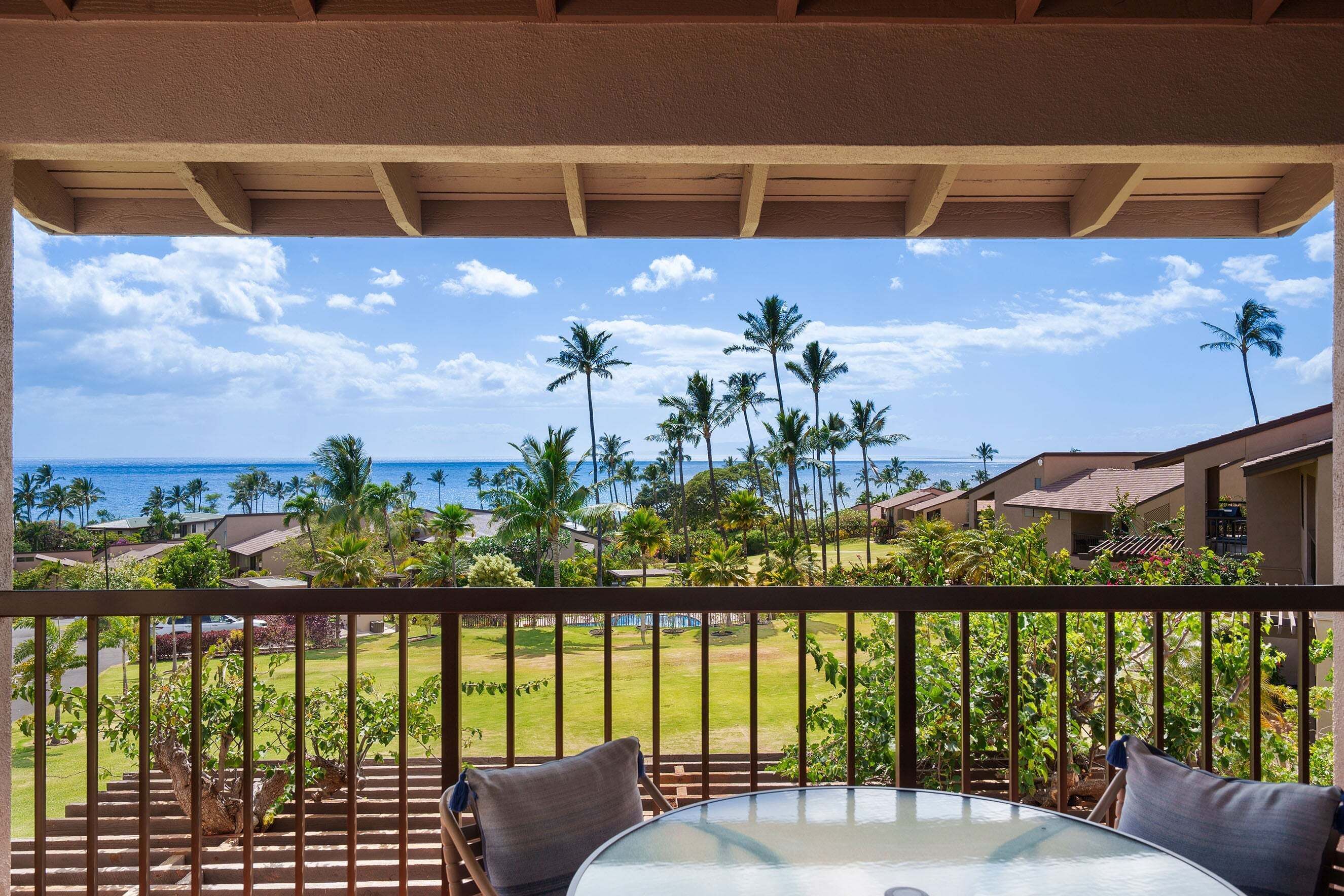 A balcony with a glass table and chairs, overlooking a tropical resort with palm trees and a view of the ocean.