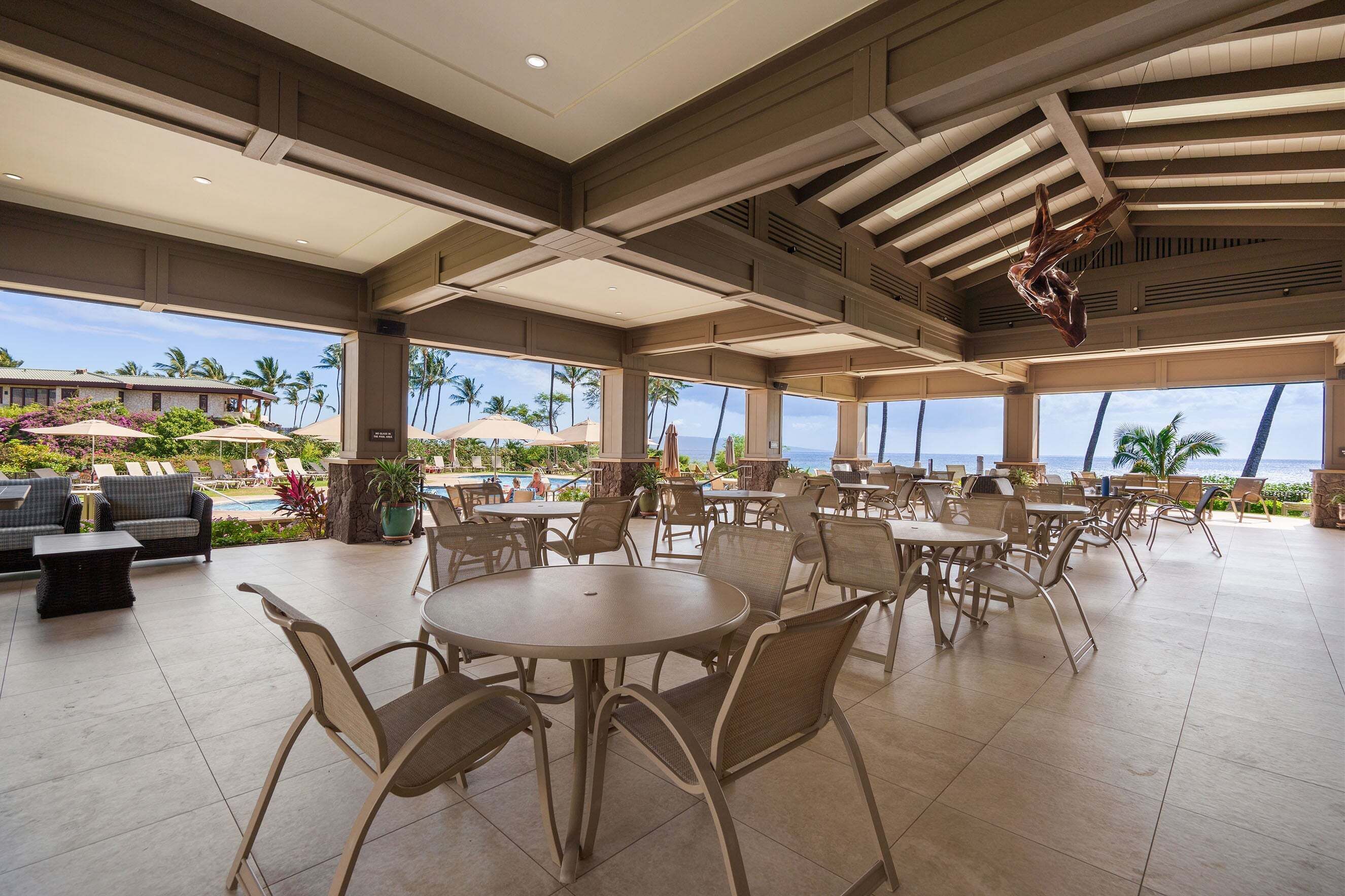 An outdoor dining area with round tables and chairs, a pool, and a view of the ocean and palm trees.