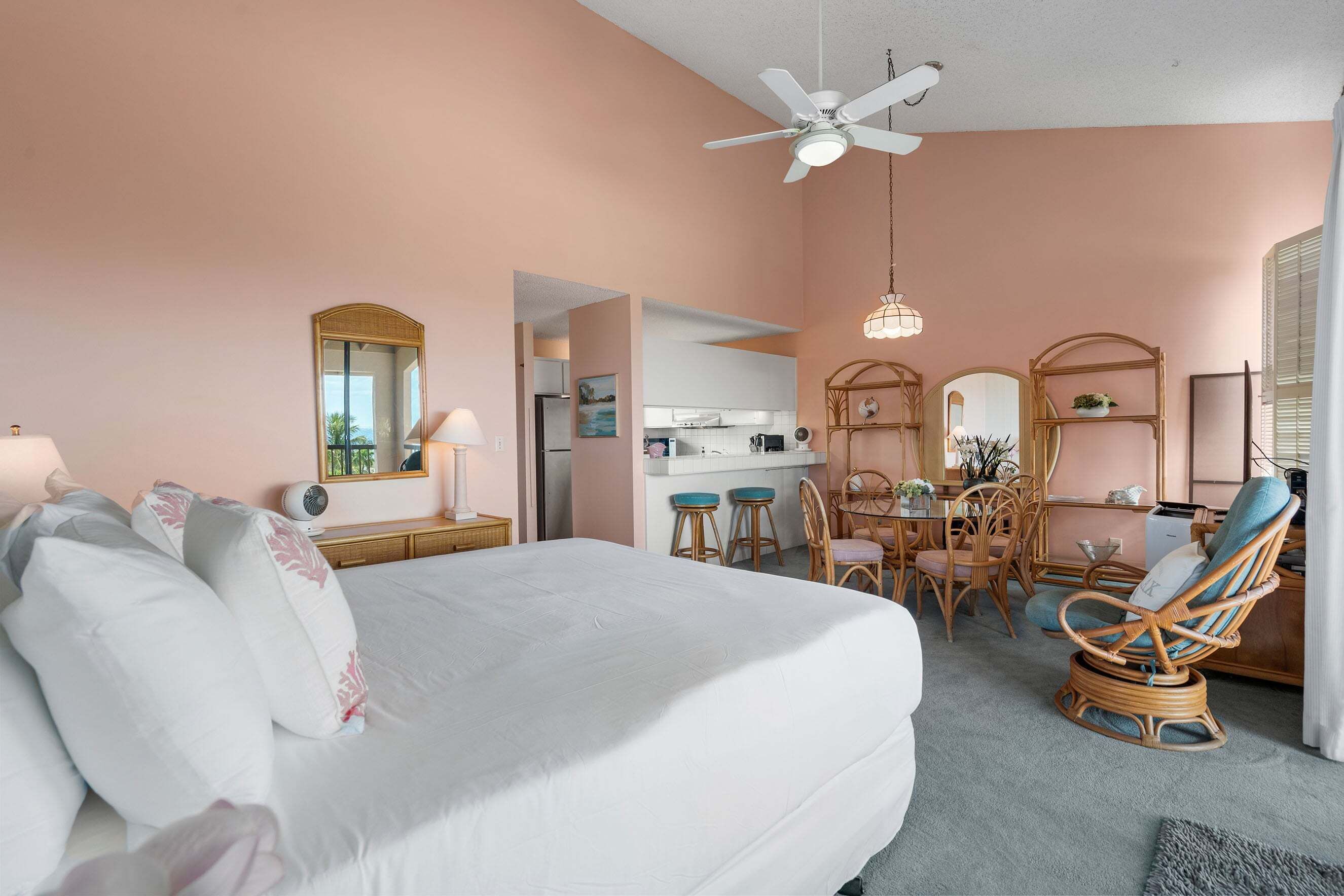 A bedroom with a bed, a ceiling fan, a dining area with chairs, and a kitchen in the background.