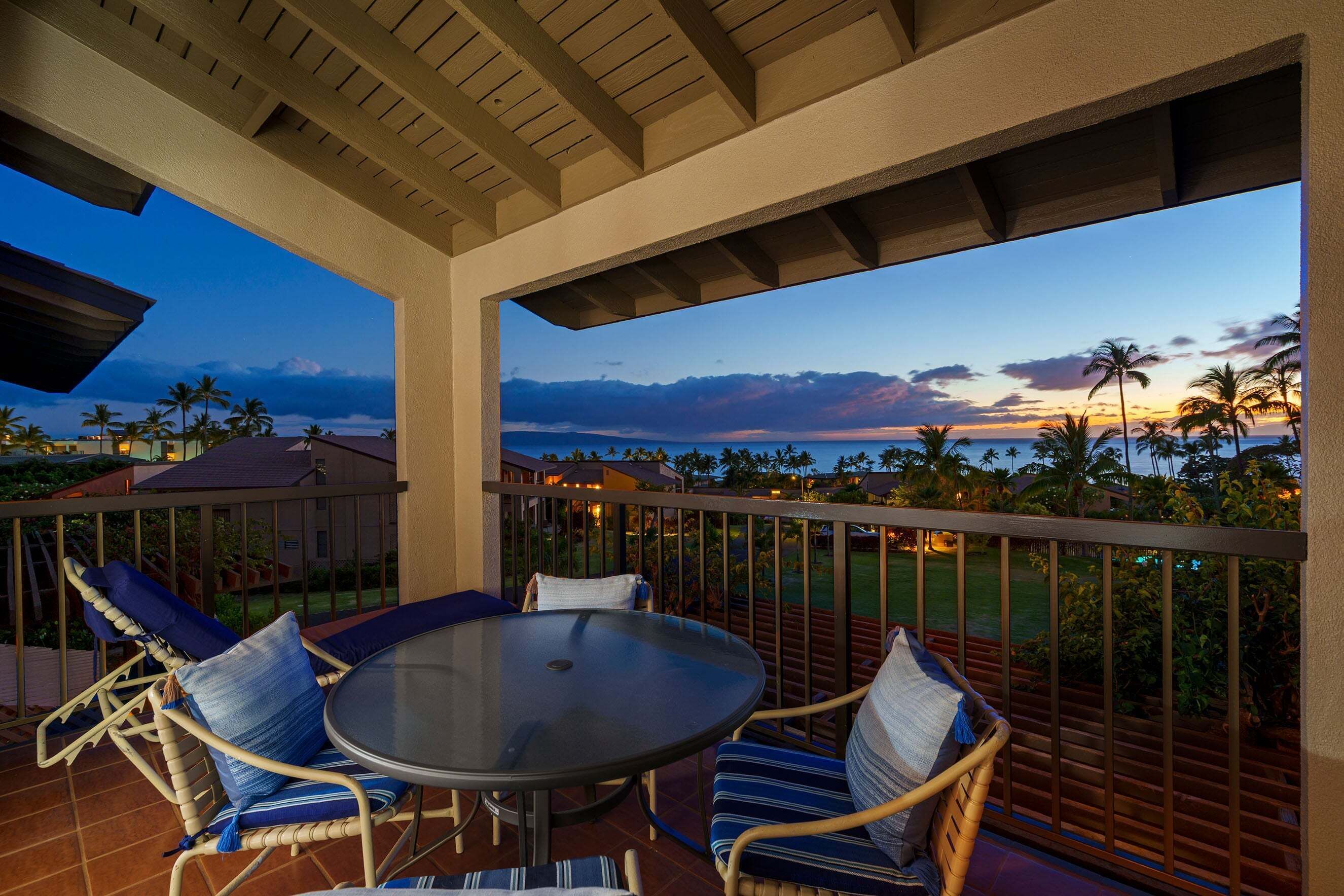 A covered outdoor seating area with a table and chairs, overlooking a scenic view of palm trees and a sunset.