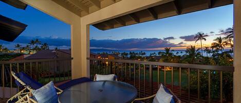 A covered outdoor seating area with a table and chairs, overlooking a scenic view of palm trees and a sunset.