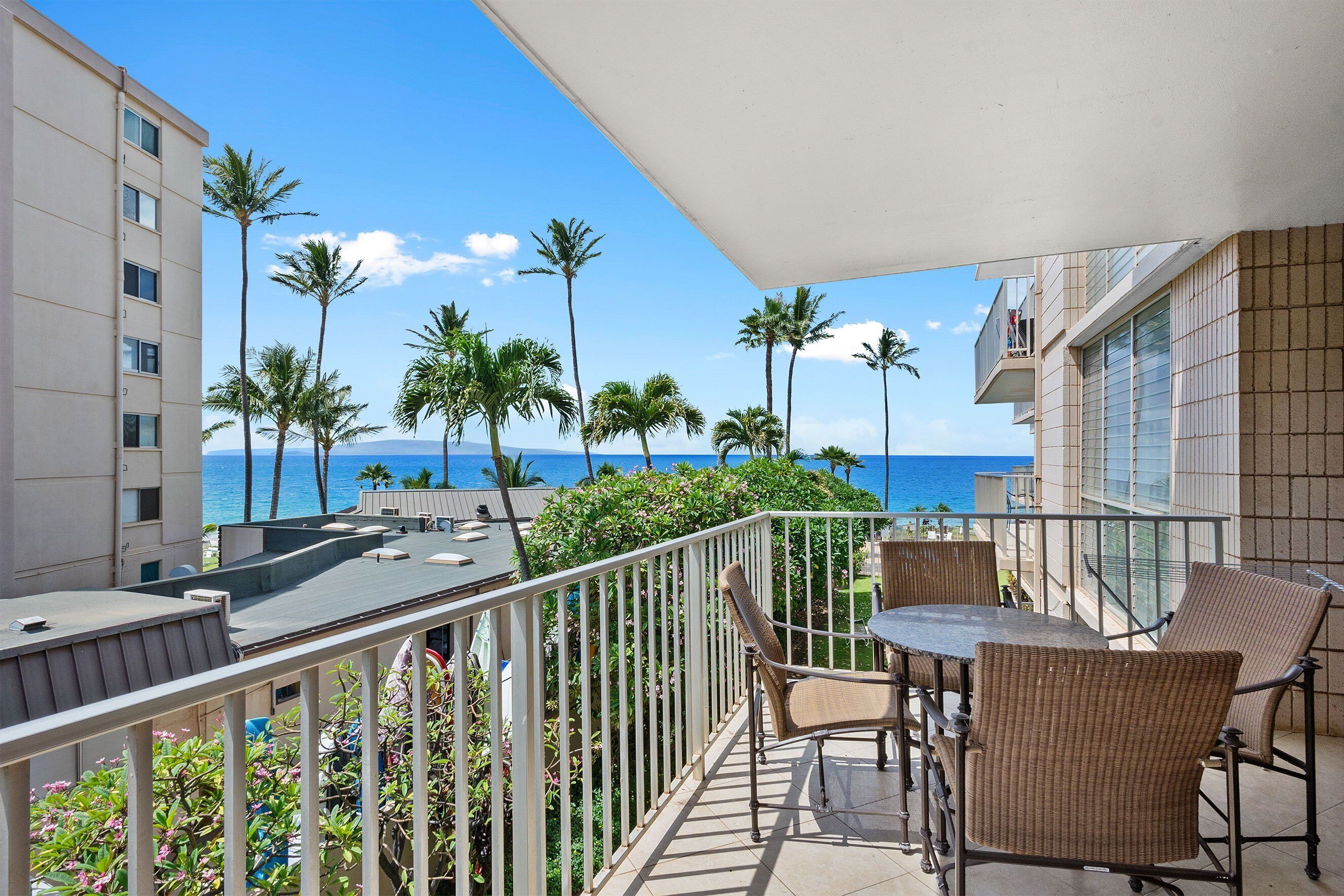 A balcony with a table and chairs, overlooking a beach with palm trees and a clear blue sky.