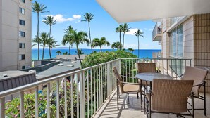 A balcony with a table and chairs, overlooking a beach with palm trees and a clear blue sky. - Kamaole Nalu 301 - Stunning Ocean Views, Renovated, Ocean Front, New Listing! (Kihei)
