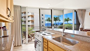 A kitchen with a large island, a stove, and a sink. There is a balcony with a view of palm trees and a building. - Kamaole Nalu 301 - Stunning Ocean Views, Renovated, Ocean Front, New Listing! (Kihei)